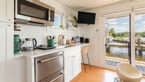 A modern kitchen with stainless steel appliances, a flat-screen TV, and a view of a water body through a sliding glass door. - Bung-a-flo Tiny Bohemian Designed Floating Home w/ 360 Waterfront Views (Arnold)