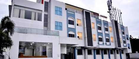 Egy modern stílusú, többemeletes szálloda, feltűnő bejárattal és jól látható, „Hotel Adarsh” felirattal ellátott táblával.