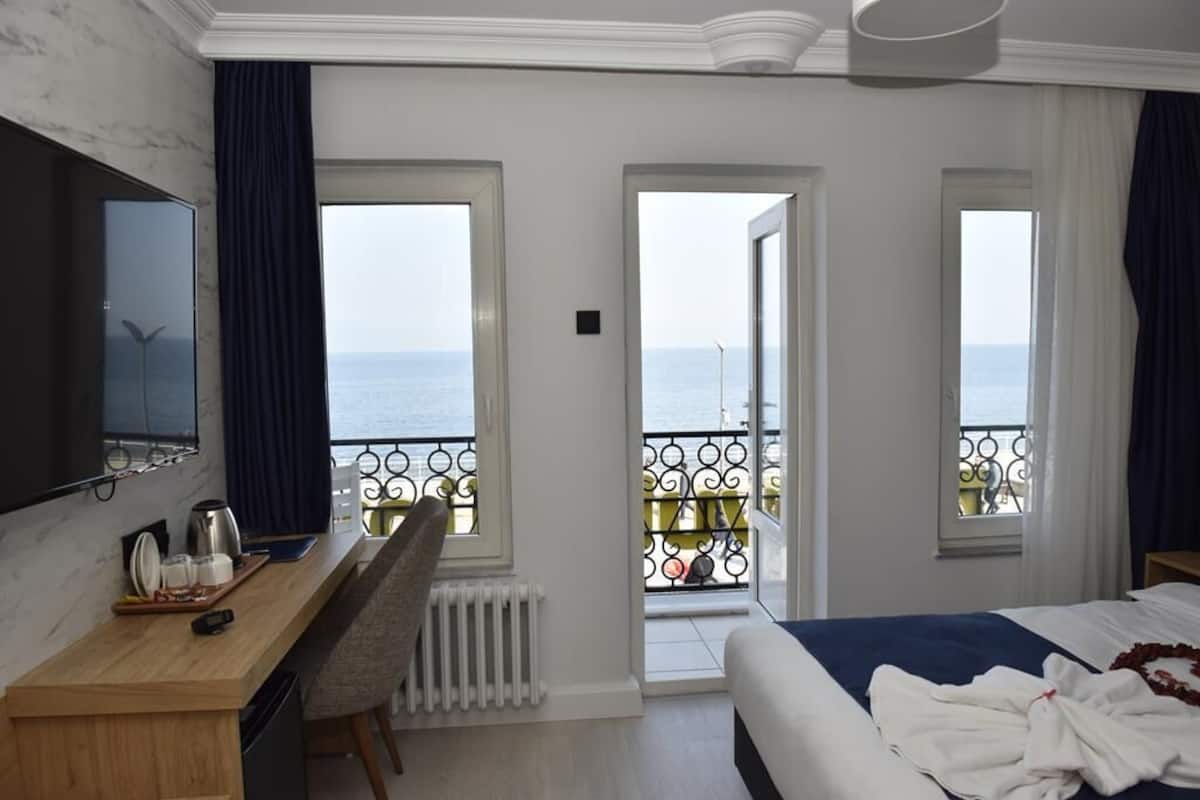 A hotel room with a bed, a desk, a chair, a TV, and a balcony with a view of the sea.