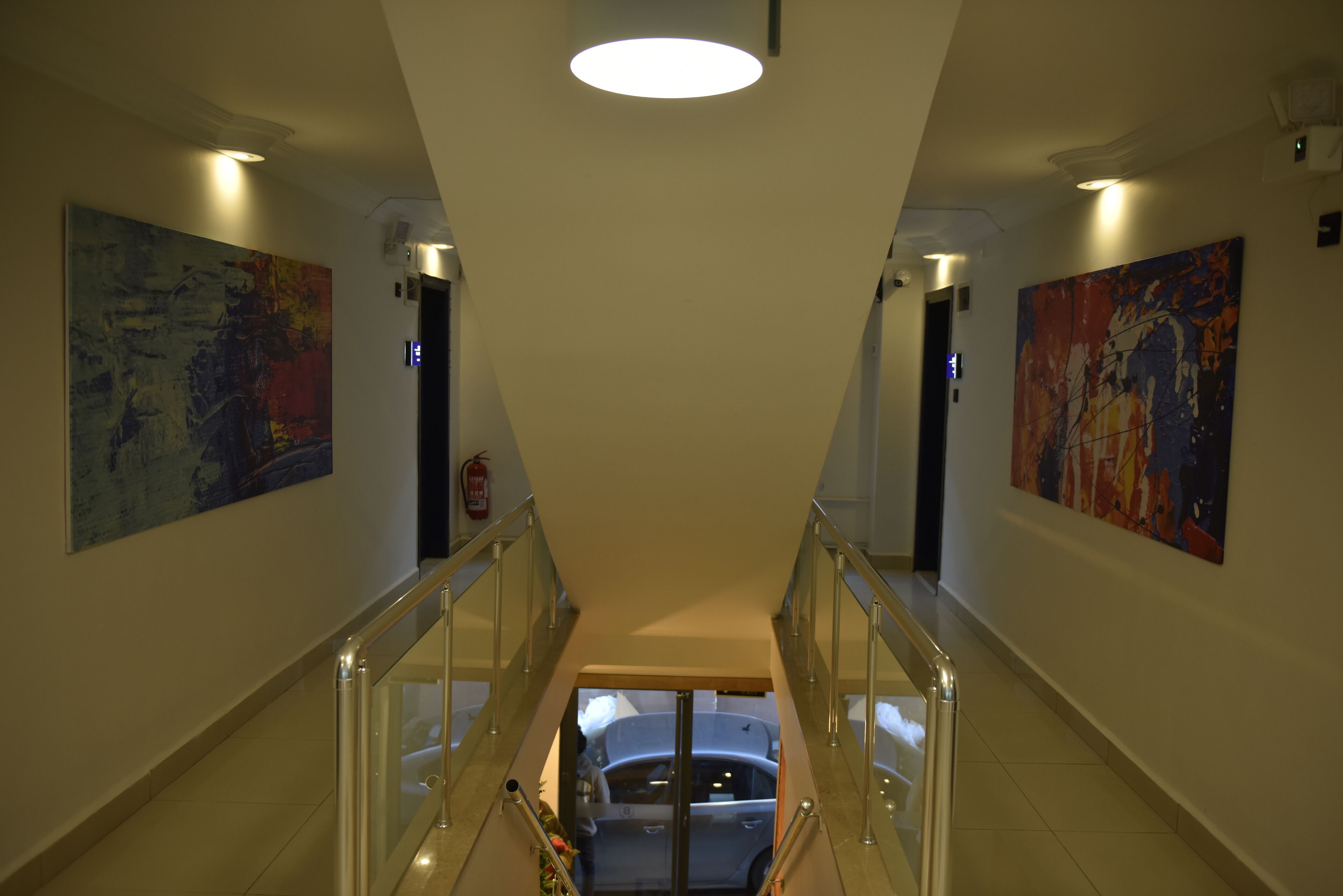 A modern hotel corridor with glass railings, artwork on the walls, and a view of a car through a glass elevator shaft.