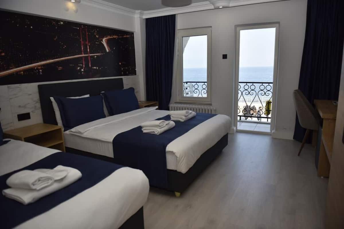 A hotel room with two beds, a large window with a view of the sea, a balcony with a railing, and a cityscape view on the wall.
