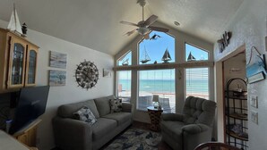 A living room with a gray sofa, a wooden chair, a TV, and a ceiling fan. There are windows with a view of the ocean and sailboats.