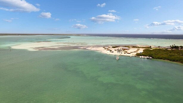 Vue aérienne d’une zone côtière avec des eaux turquoise limpides, une plage de sable et un petit village avec des bateaux et des bâtiments.