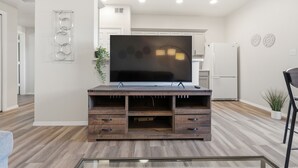 A modern living room with a flat-screen TV on a wooden entertainment center, a kitchen in the background, and a dining area with a chair. - Renovated 3BR with Yard & Mountain Views (Rio Rancho)