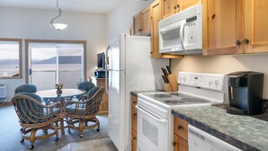 A kitchen with wooden cabinets, a white stove, a microwave, and a coffee maker. There is a dining table with chairs and a view of the outdoors through a large window. - Lakefront Studio with views! (Bigfork)