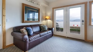 A room with a brown leather sofa, a framed picture on the wall, a floor lamp, and a view through a glass door to the outside. - New! Waterfront Studio near Glacier! (Bigfork)