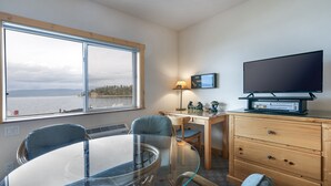 A room with a large window, a glass table, two chairs, a wooden desk, a television, and a lamp. - New! Waterfront Studio near Glacier! (Bigfork)