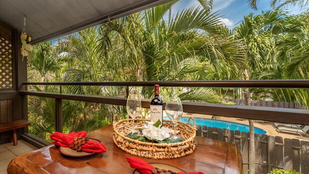 A dining table with a bottle of wine, two glasses, and a menu on a balcony overlooking a pool and palm trees.