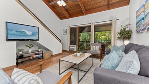 A modern living room with a flat-screen TV, wooden ceiling beams, a ceiling fan, a sofa, a coffee table, and a sliding glass door leading to a balcony.