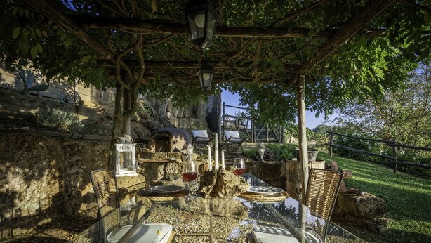 Ein Essbereich im Freien mit Tischen und Stühlen, einer Pergola und einer Steinmauer im Hintergrund.