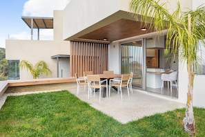 A modern outdoor dining area with a wooden table and chairs, a grassy lawn, and a view of the house.