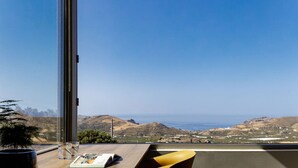 A room with a large window overlooking a coastal landscape, a wooden table with a magazine, and a yellow chair. - Seaview villa w/private pool, 4km to Plakias beach (Mariou)
