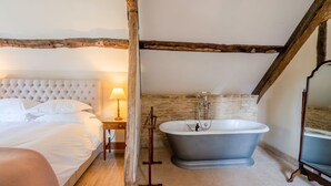 A bedroom with a large bed, a nightstand with a lamp, and a stone wall. - The Coach House - sleeps 4 guests  in 2 bedrooms (Cold Aston)