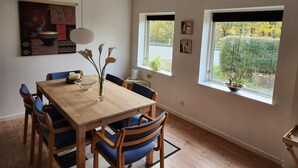 A dining area with a wooden table and blue chairs, a vase with flowers, and two windows with views of greenery. - Breakaway Cottage (Grasten)