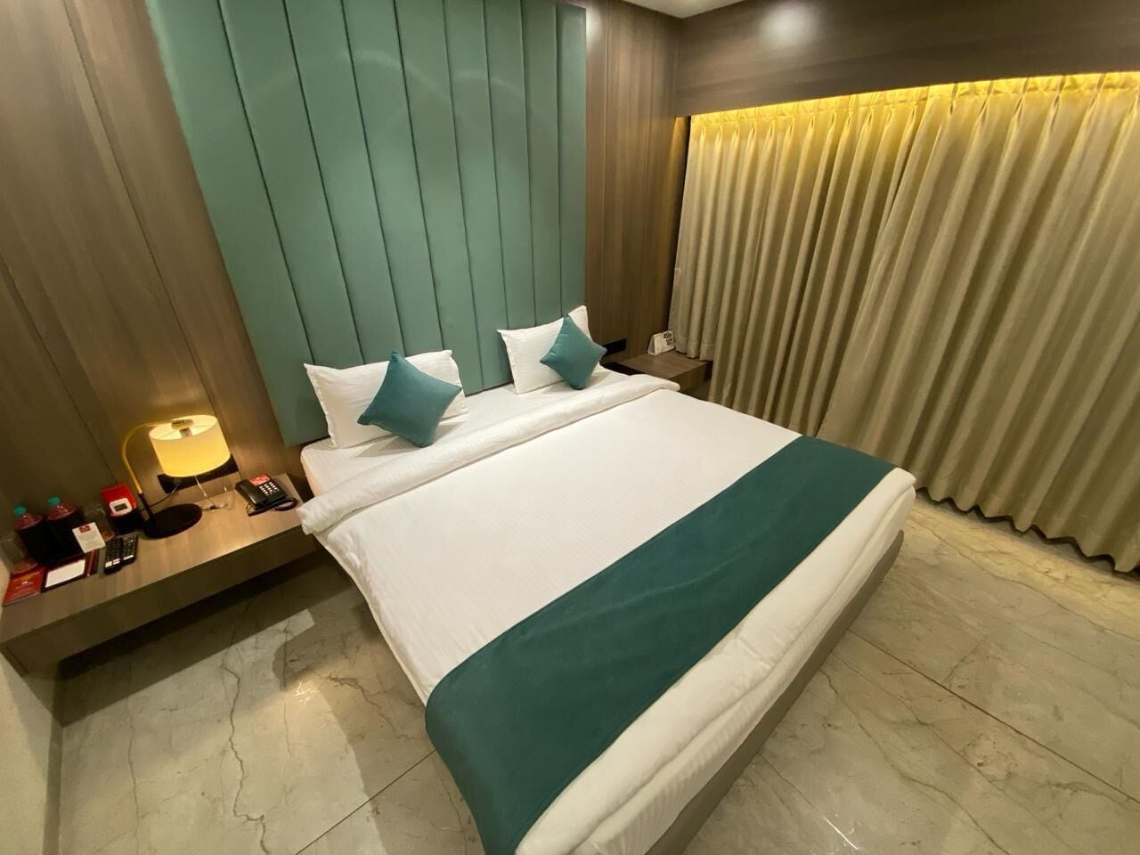 Habitación de hotel con una cama grande, una mesita de noche con una lámpara, un teléfono y una pequeña mesa con un libro y una botella.
