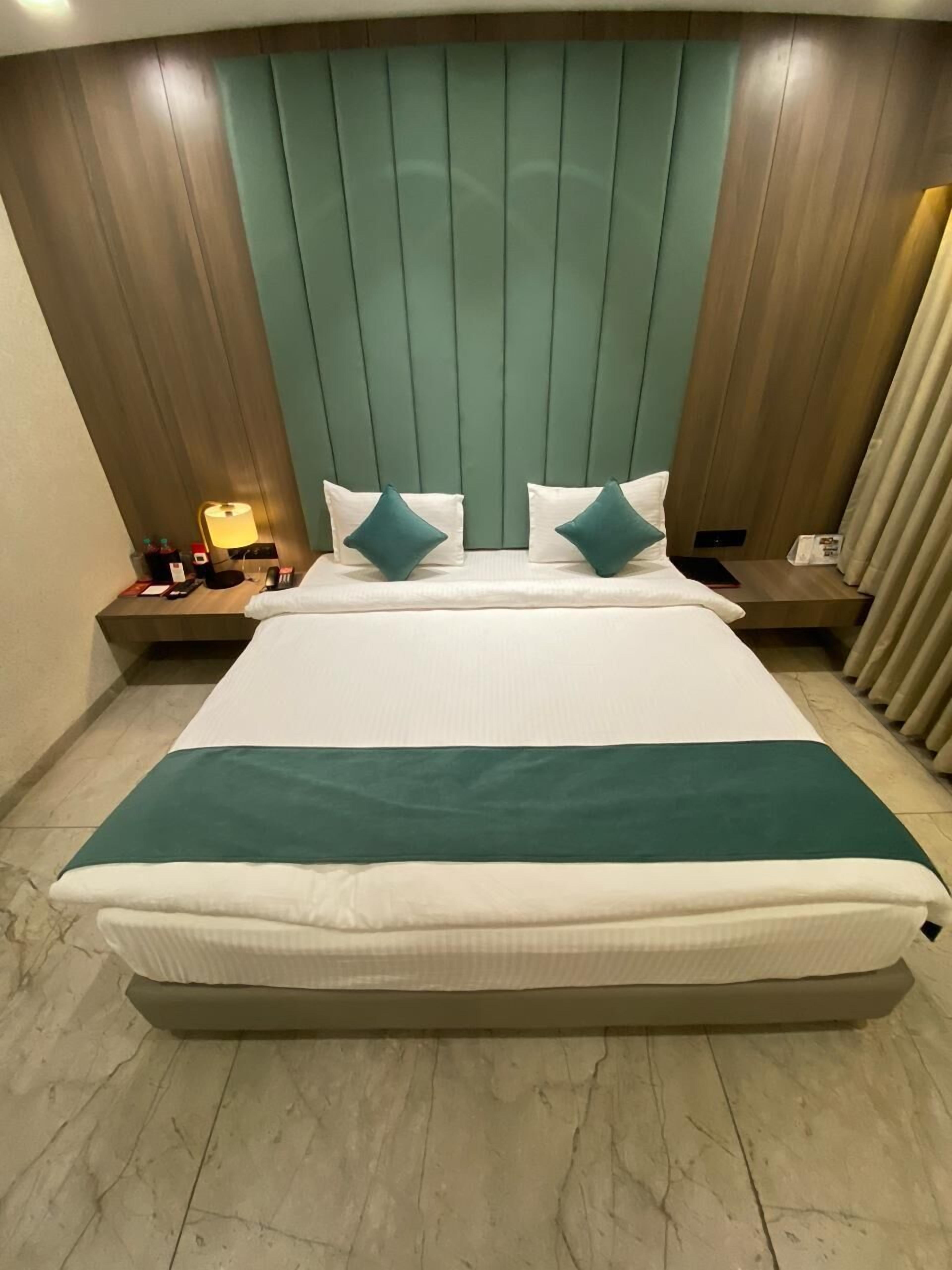 A hotel room with a large bed, two pillows, a bedside table with a lamp, and a nightstand with a phone and other items.