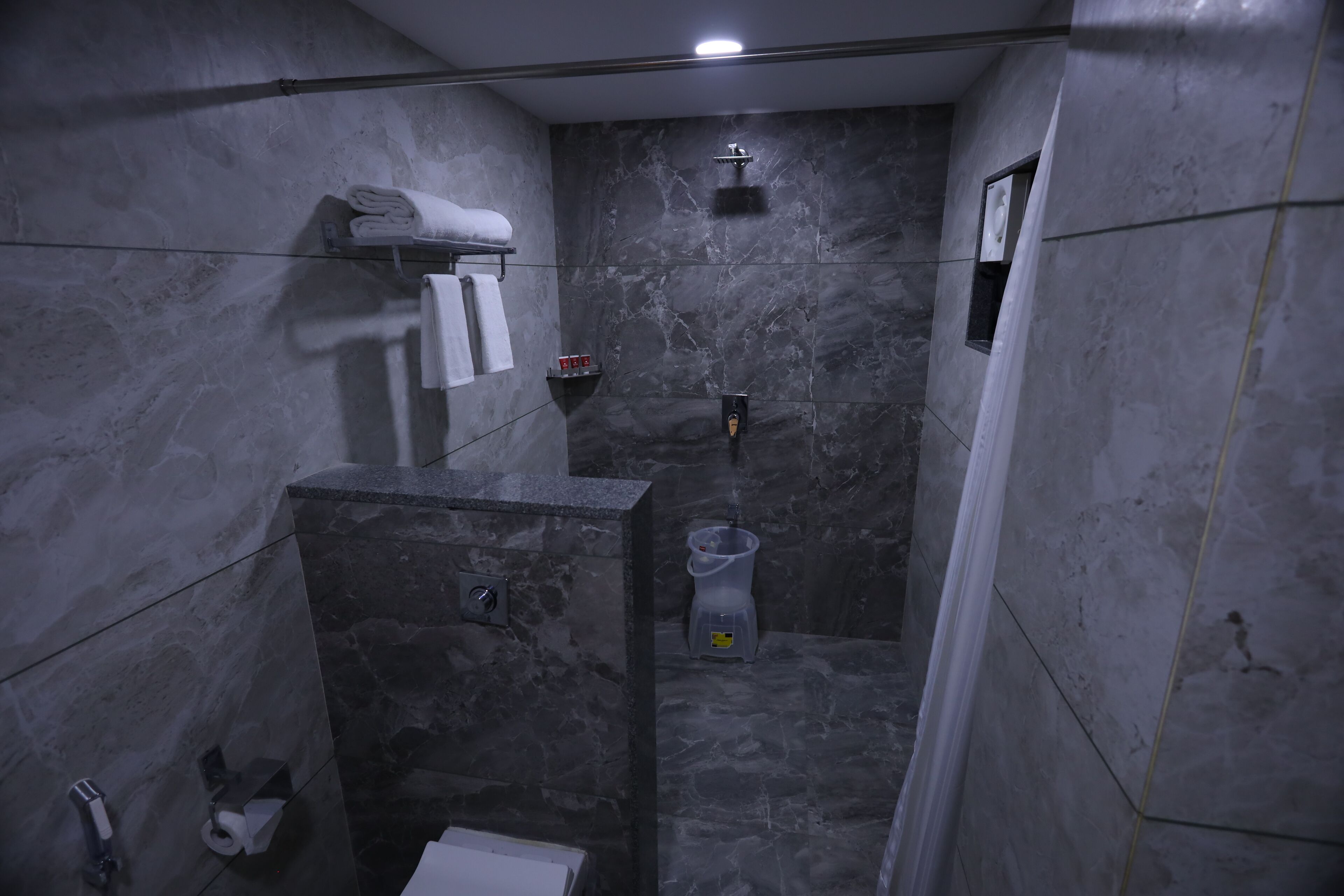 A narrow, marble-floored shower area with a handheld showerhead, a towel rack with folded towels, and a small shelf with toiletries.