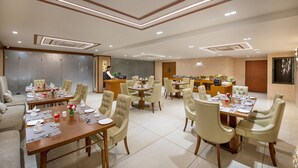 A hotel dining area with multiple tables set for guests, a reception desk, and a kitchen area in the background.