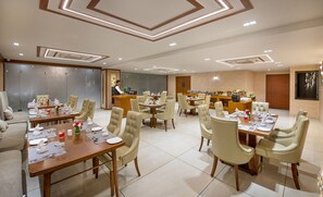 A hotel dining area with multiple tables set for guests, a reception desk, and a kitchen area in the background.
