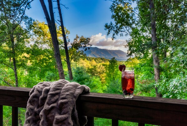 Un verre de thé glacé, décoré d’une fraise, posé sur une terrasse en bois donnant sur une zone boisée, avec des montagnes visibles au loin.