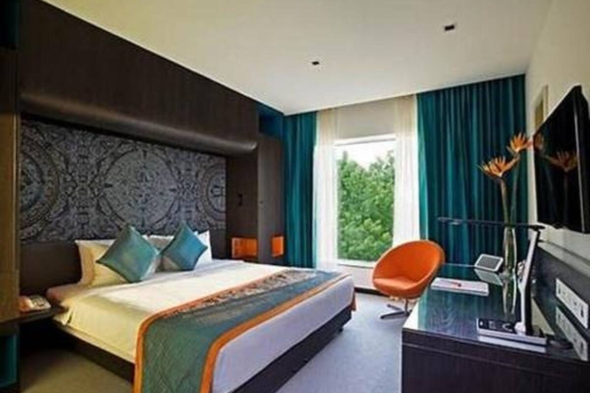 A modern hotel room with a large bed, a desk with a laptop, and a view of greenery outside.