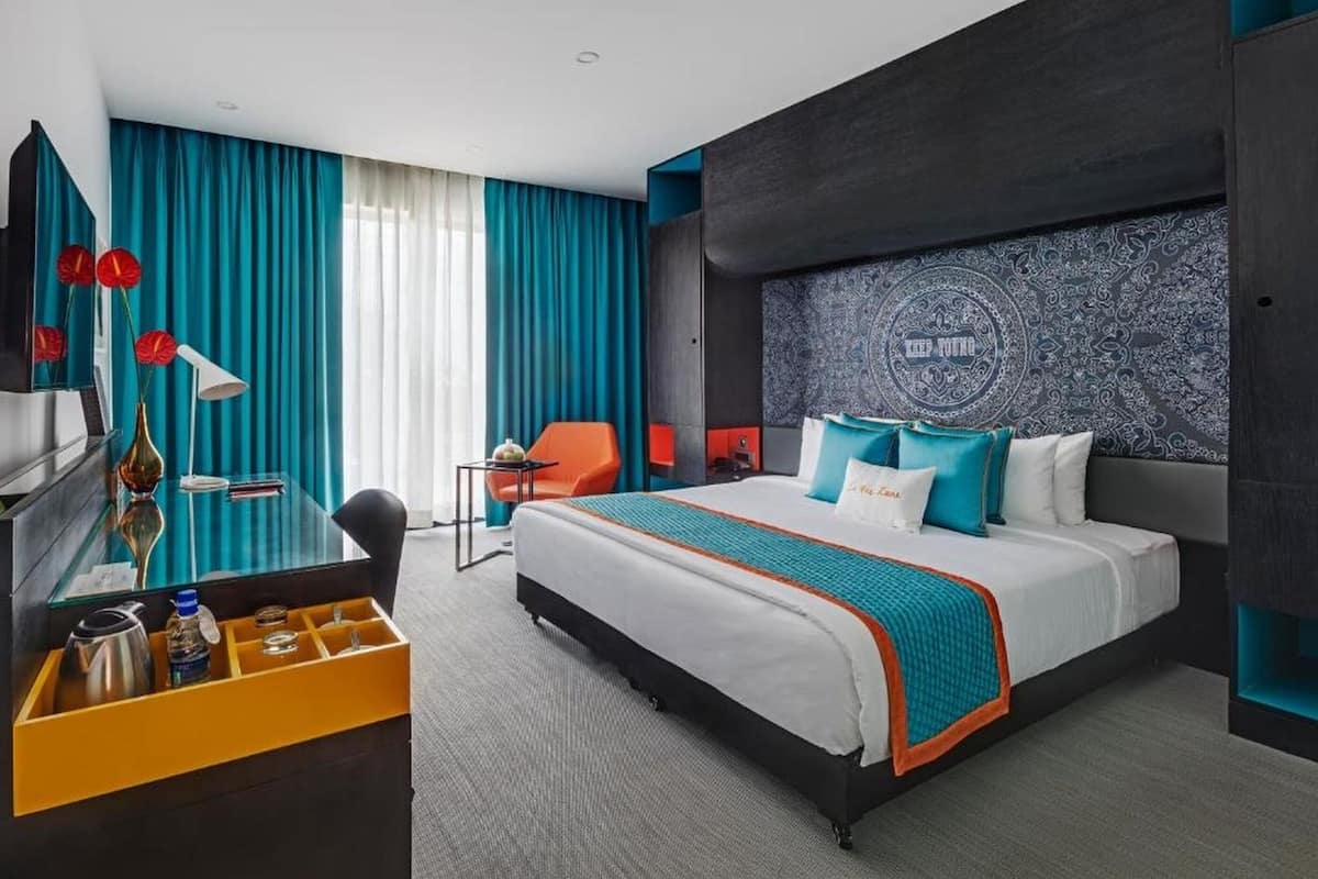 A modern hotel room with a large bed, a desk with a chair, a small table, and a minibar.
