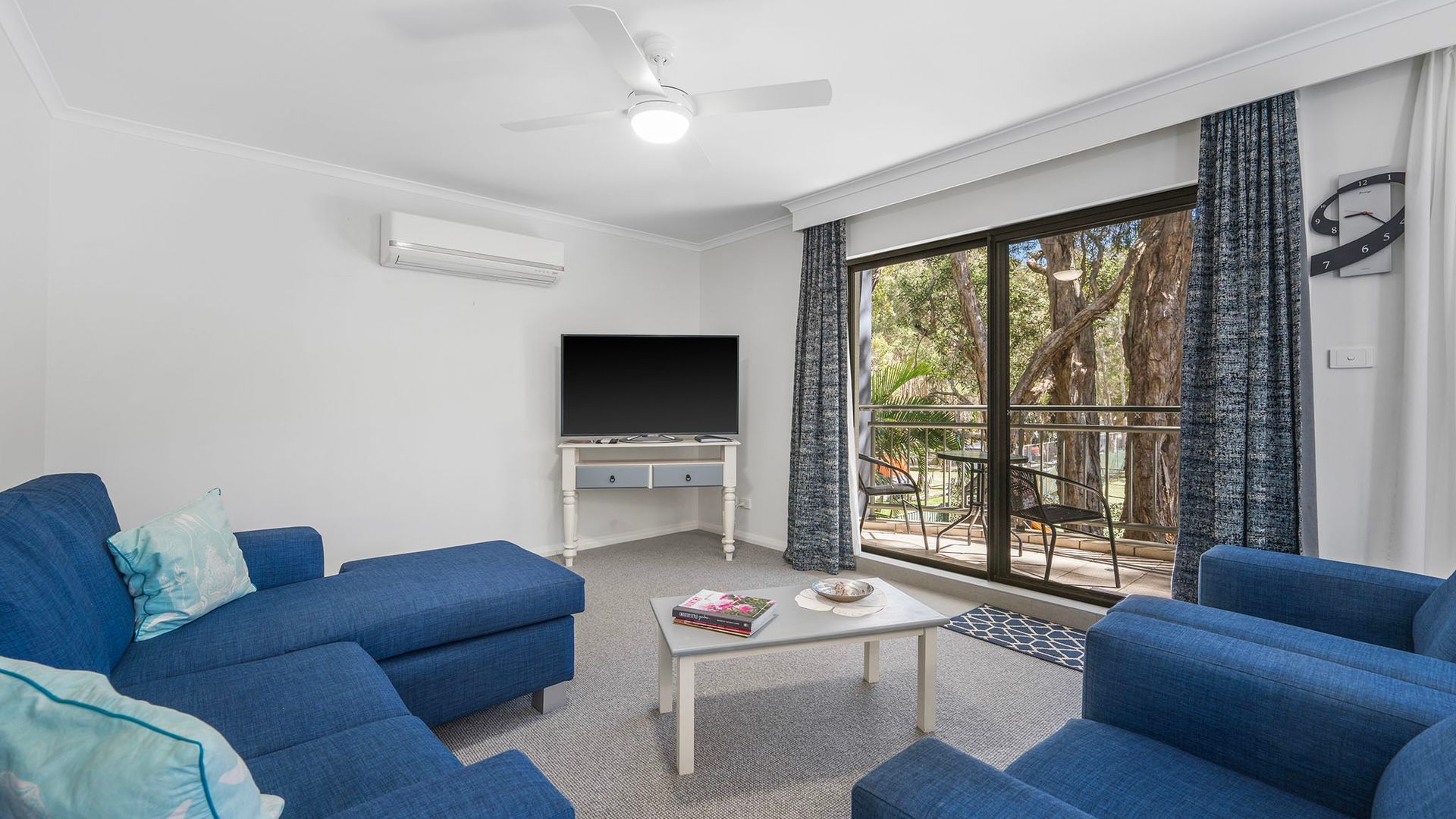 Ein Wohnzimmer mit Flachbildfernseher, blauen Sofas, einem Couchtisch mit Büchern und einem Balkon mit Vorhängen.