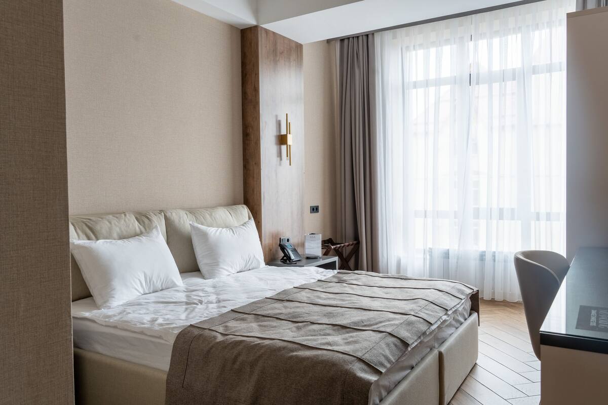 A modern hotel room with a large bed, a bedside table with a lamp, a chair, and a large window with sheer curtains.