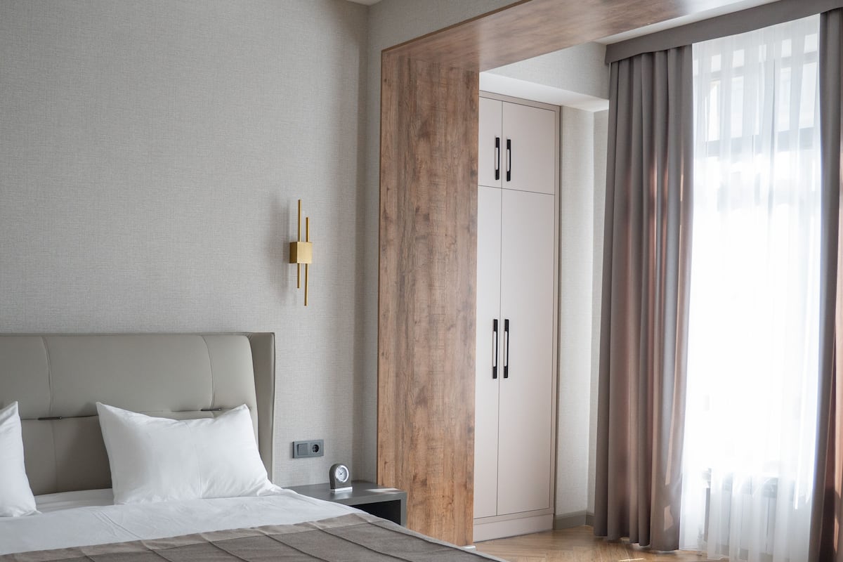A modern bedroom with a large bed, a nightstand, a wall-mounted light, a wardrobe, and a window with curtains.