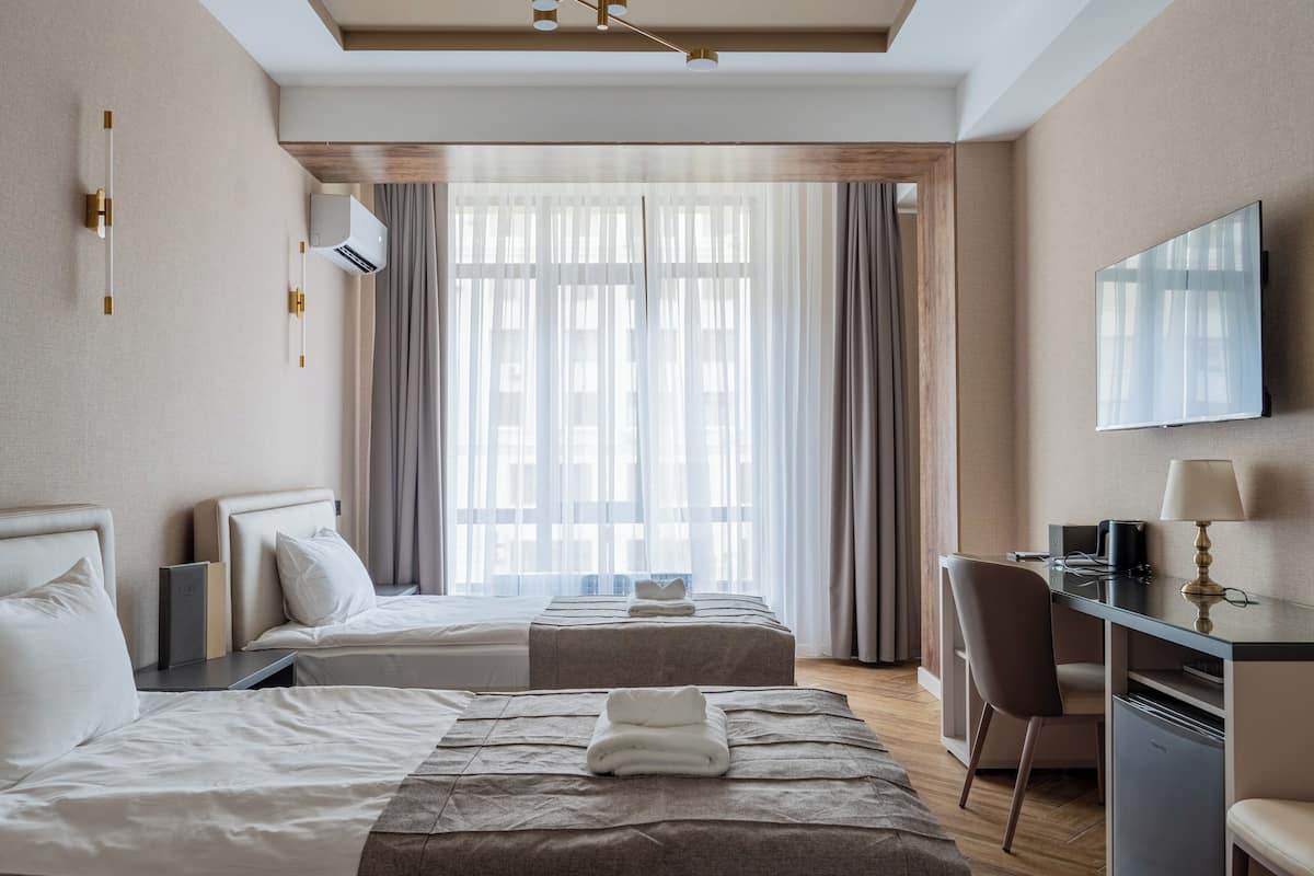 A hotel room with two beds, a desk, a chair, a TV, and a large window with curtains.