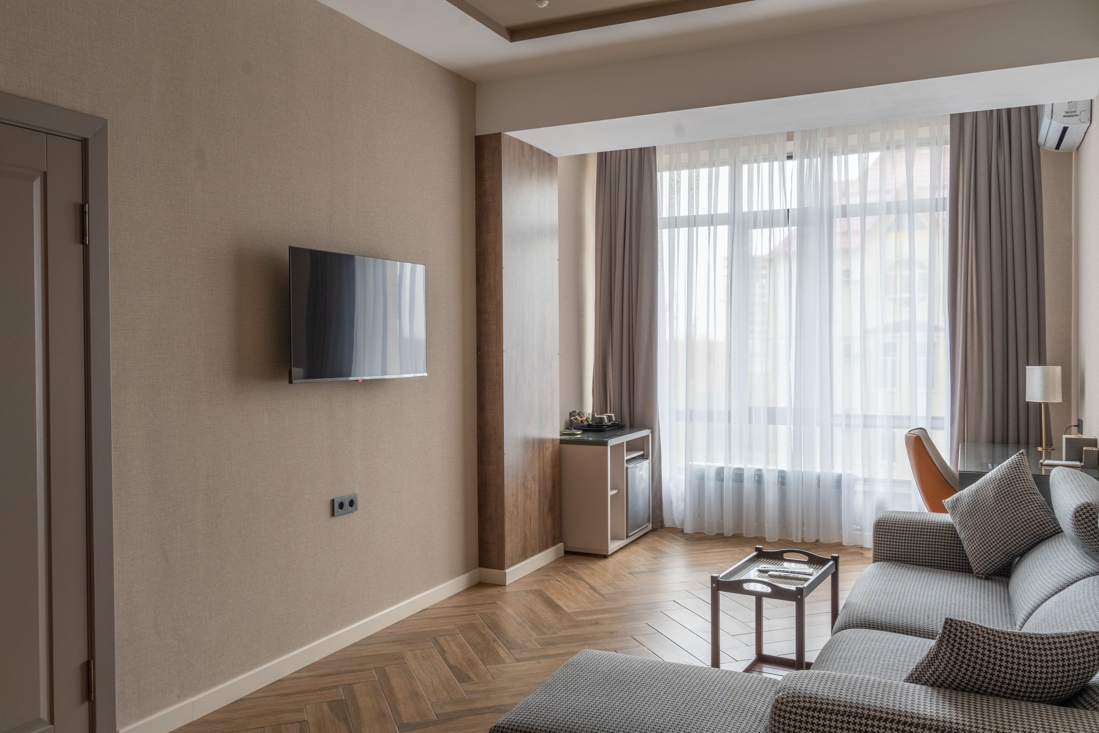 Una sala moderna con un televisor de pantalla plana montado en la pared, un juego de sofá, una mesita y una ventana con cortinas traslúcidas.