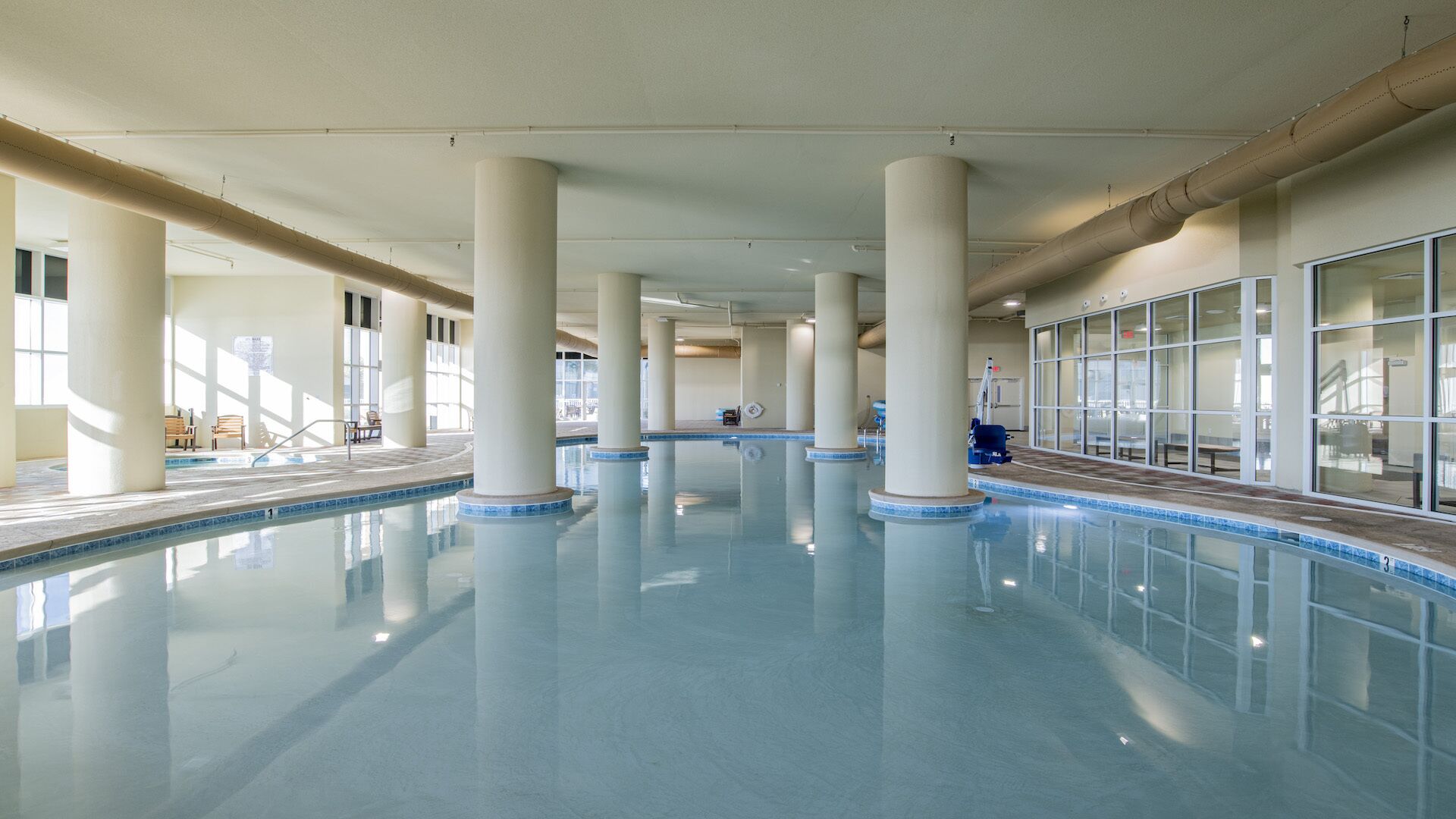 Une vaste piscine intérieure à l’eau d’un bleu limpide, entourée de colonnes blanches et de murs de verre.