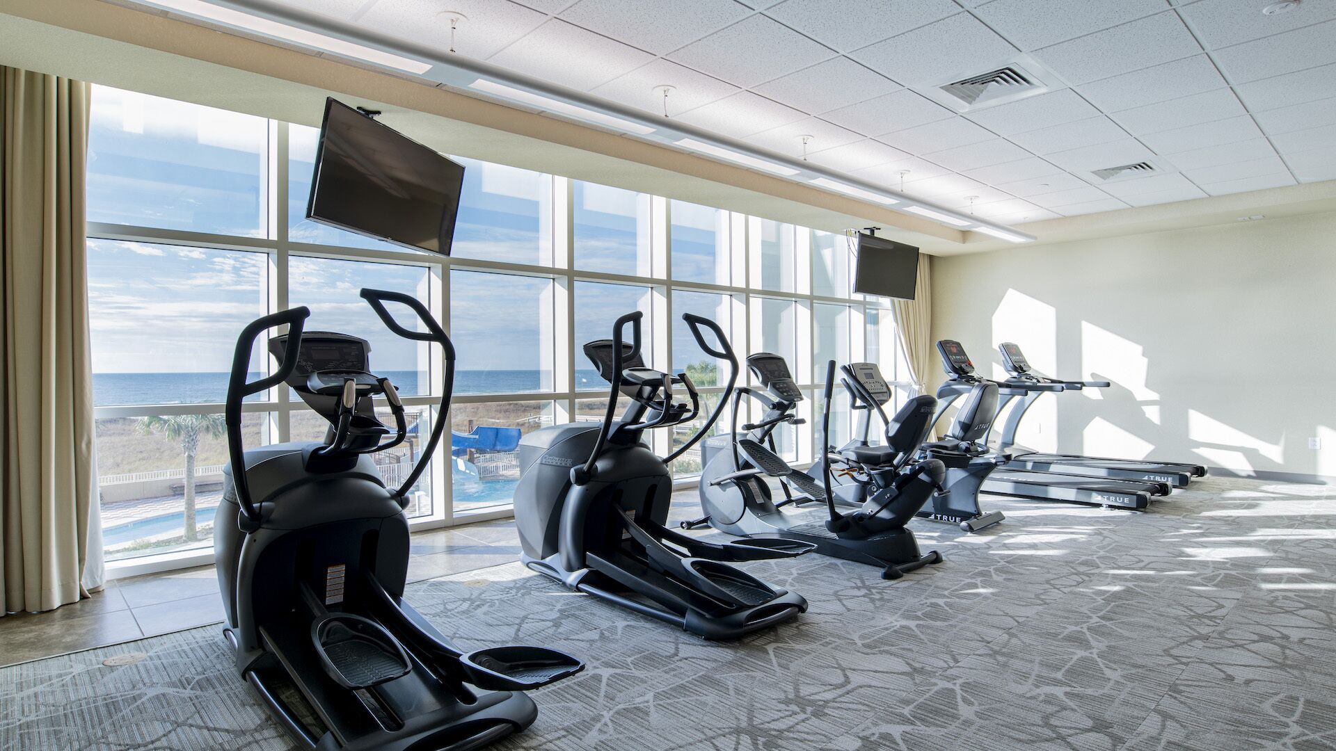 A gym with multiple exercise machines, large windows with a view of the ocean, and a ceiling with recessed lighting.