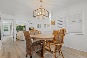 Een eetgedeelte met een ronde tafel, vier rieten stoelen, een hanglamp en witte muren met zonwering.