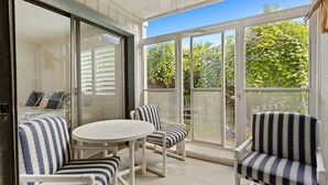 A covered patio with a white table and striped chairs, a sliding glass door, and a view of greenery outside. - Beachside Gem!  Sea Woods Condo 153 - Close to Pool & Beach Access (New Smyrna Beach)