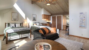 Una habitación amplia con techo alto, una cama con ropa de cama estampada, un sofá con cojines, una mesa de comedor con una cesta, y una zona de cocina con armarios de madera.