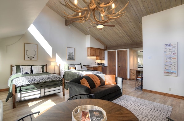Une pièce spacieuse avec un plafond haut, un lit avec une literie à motifs, un canapé avec des coussins, une table à manger avec un panier, et un coin cuisine avec des placards en bois.