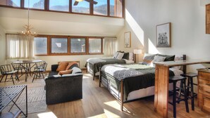 Amplia habitación con techo alto, ventanas grandes, ventilador de techo, zona de comedor con mesa y sillas, dos camas, una pequeña kitchenette y un escritorio de madera con taburetes.