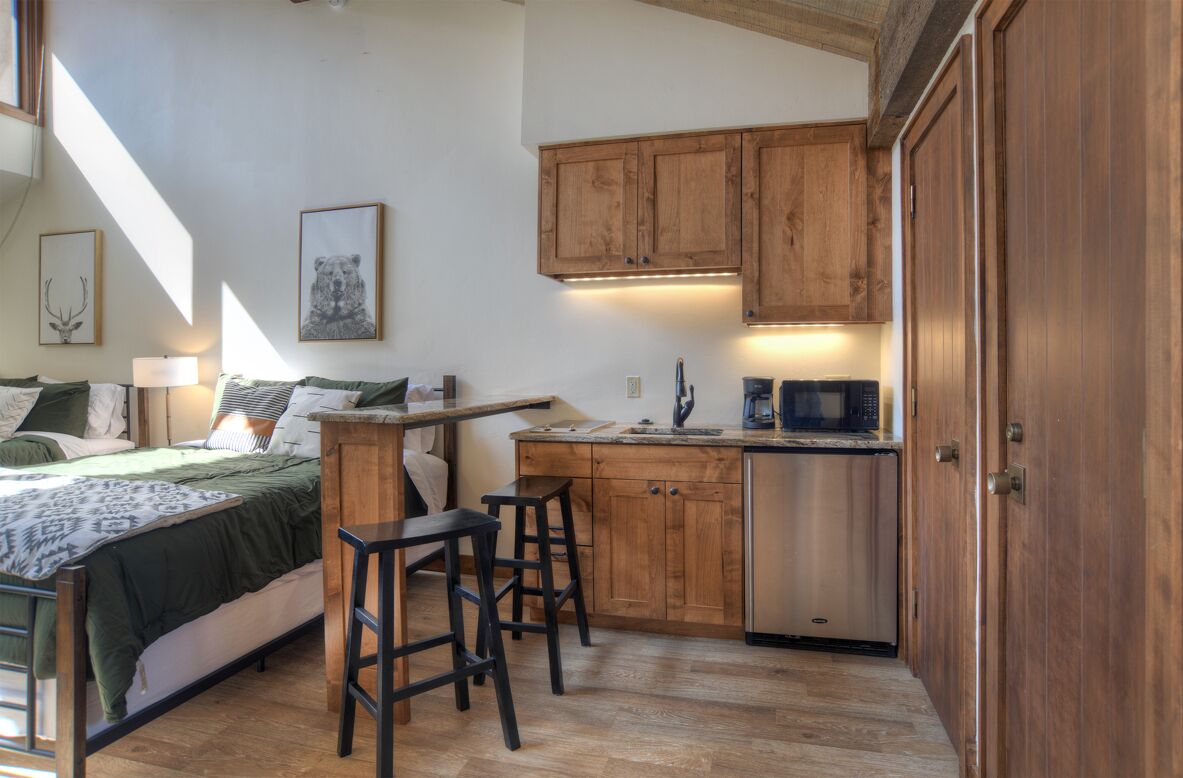 A compact bedroom with a bed, bedside table, and a small kitchenette area with a microwave and wooden cabinets.