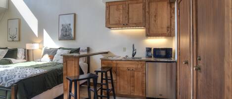 A compact bedroom with a bed, bedside table, and a small kitchenette area with a microwave and wooden cabinets.