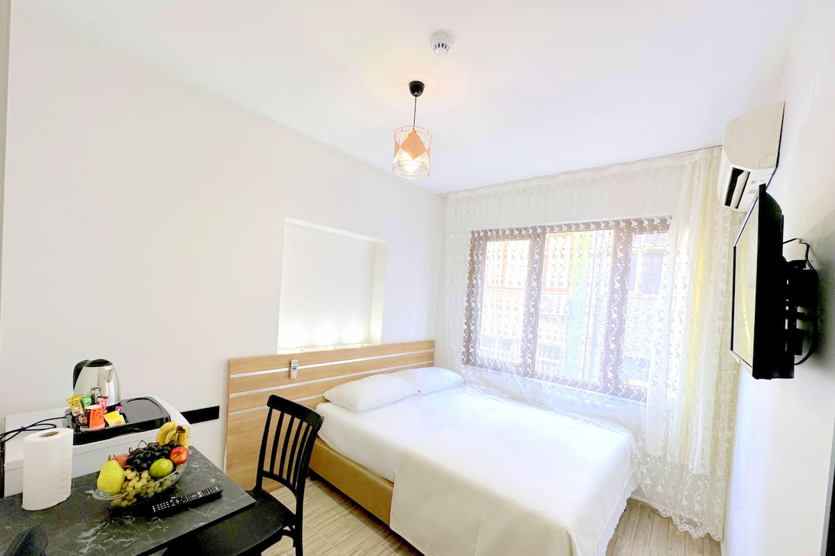 A hotel room with a bed, a desk with a fruit bowl, a chair, a table, a kettle, a remote control, and a TV mounted on the wall.