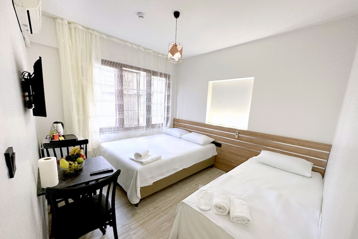 A hotel room with two beds, a desk with a chair, a small table with a fruit bowl, and a window with sheer curtains.