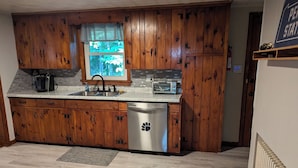 A kitchen with wooden cabinets, a stainless steel dishwasher, a window with a view of trees, and a countertop with a coffee maker and toaster. - 2BR Sleeps 8 near Penn State, Stadium & Downtown with Hot Tub (State College)