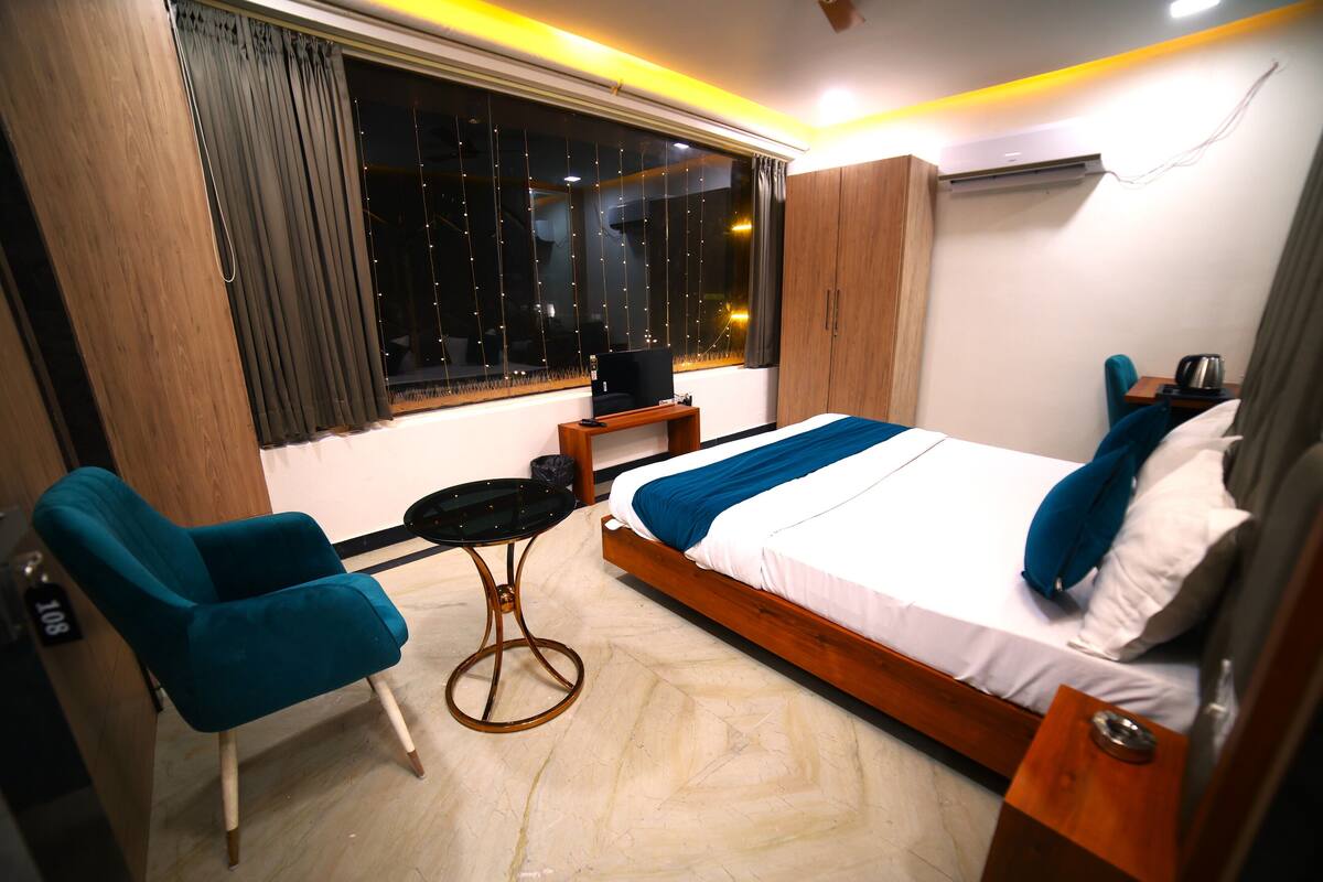 Una habitación de hotel moderna con una cama grande, una mesita pequeña, un sillón azul y un ventanal con cortinas.