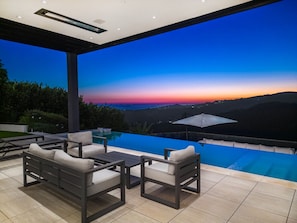 A modern outdoor lounge area with a pool, seating, and a view of a sunset over a hilly landscape.