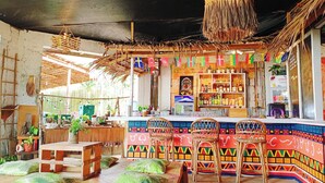 Eine lebhafte Bar mit einer farbenfrohen Wandmalerei, Holztischen und Regalen voller Flaschen und Snacks.