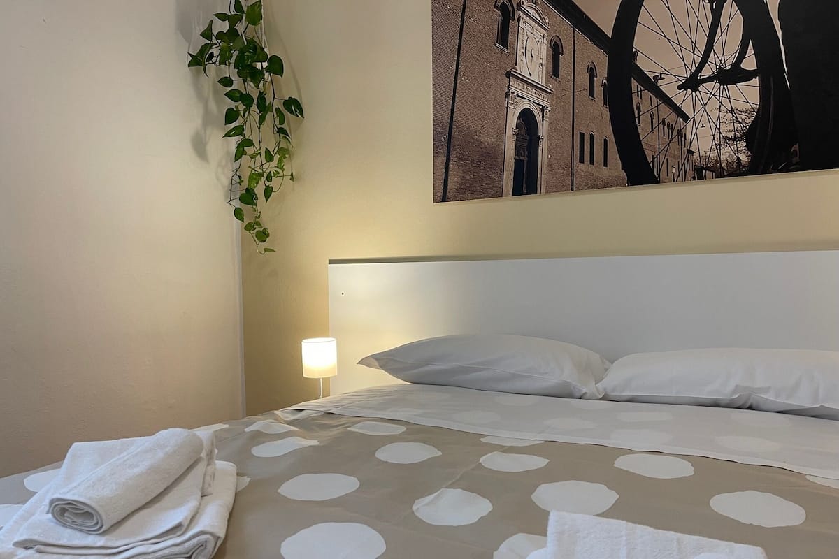 A hotel room with a bed, a nightstand with a lamp, a wall-mounted plant, and a large painting of a building.