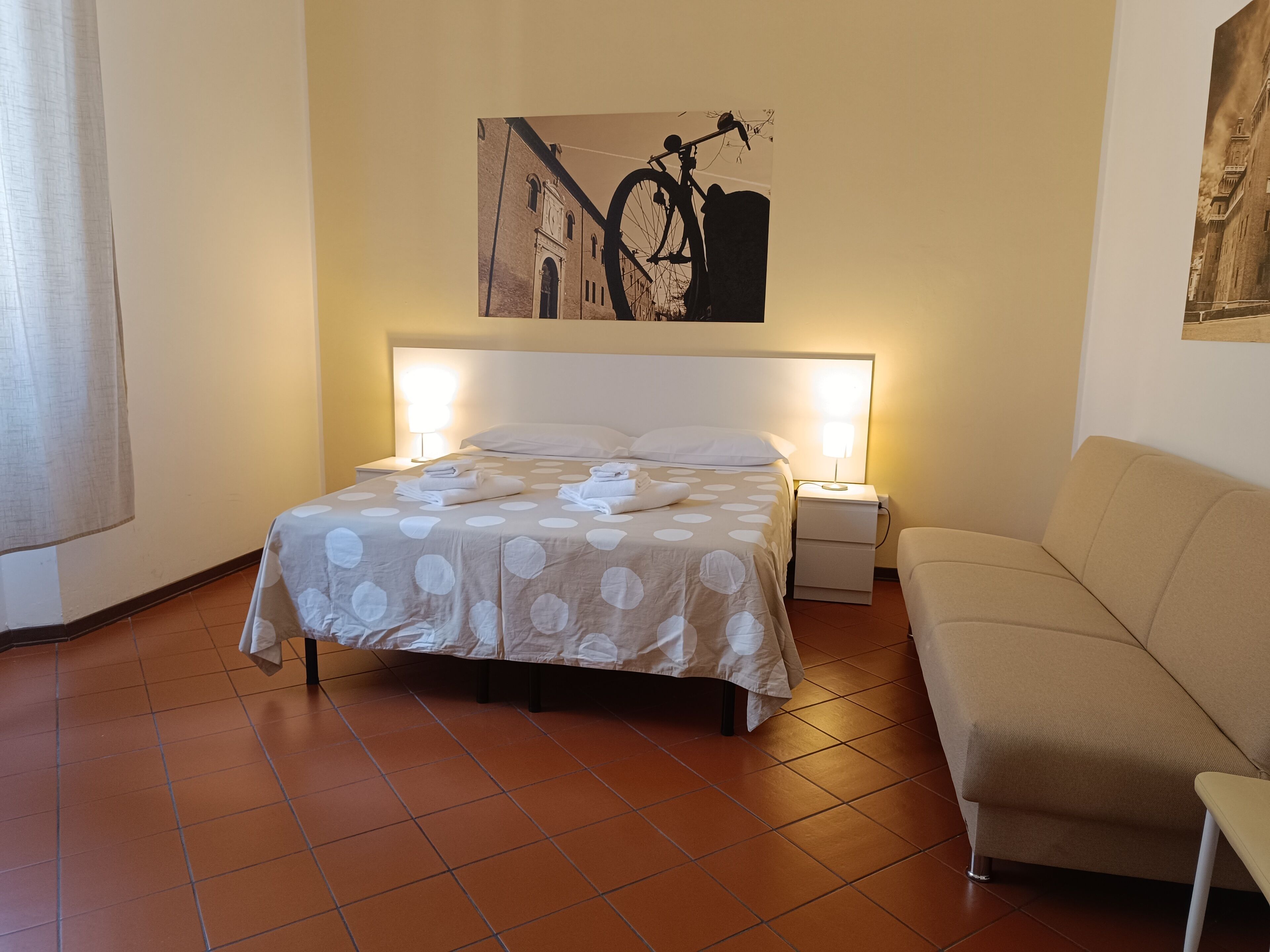A hotel room with a bed, a nightstand, a lamp, a sofa, and a painting on the wall.