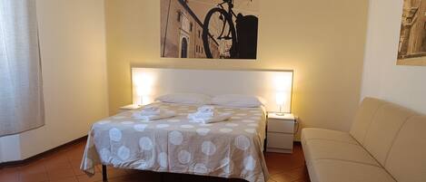 A hotel room with a bed, a nightstand, a lamp, a sofa, and a painting on the wall.