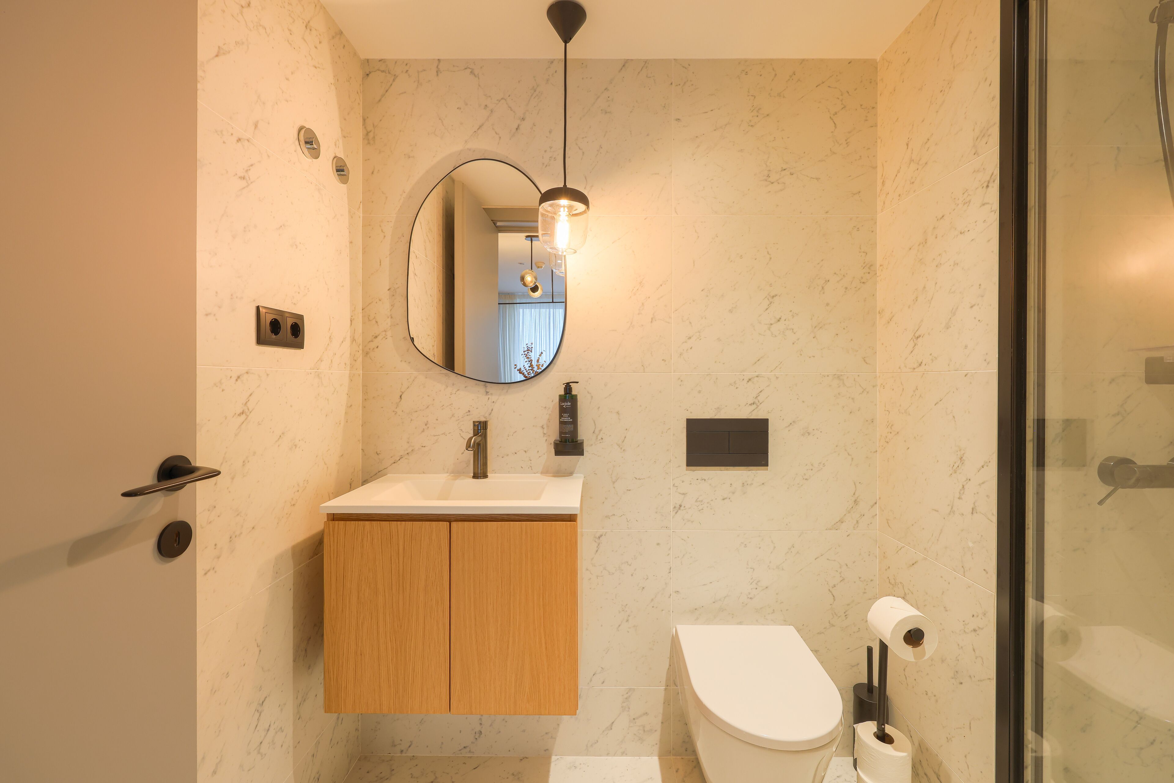 A modern bathroom with a marble wall, a wooden vanity, a round mirror, and a glass shower enclosure.